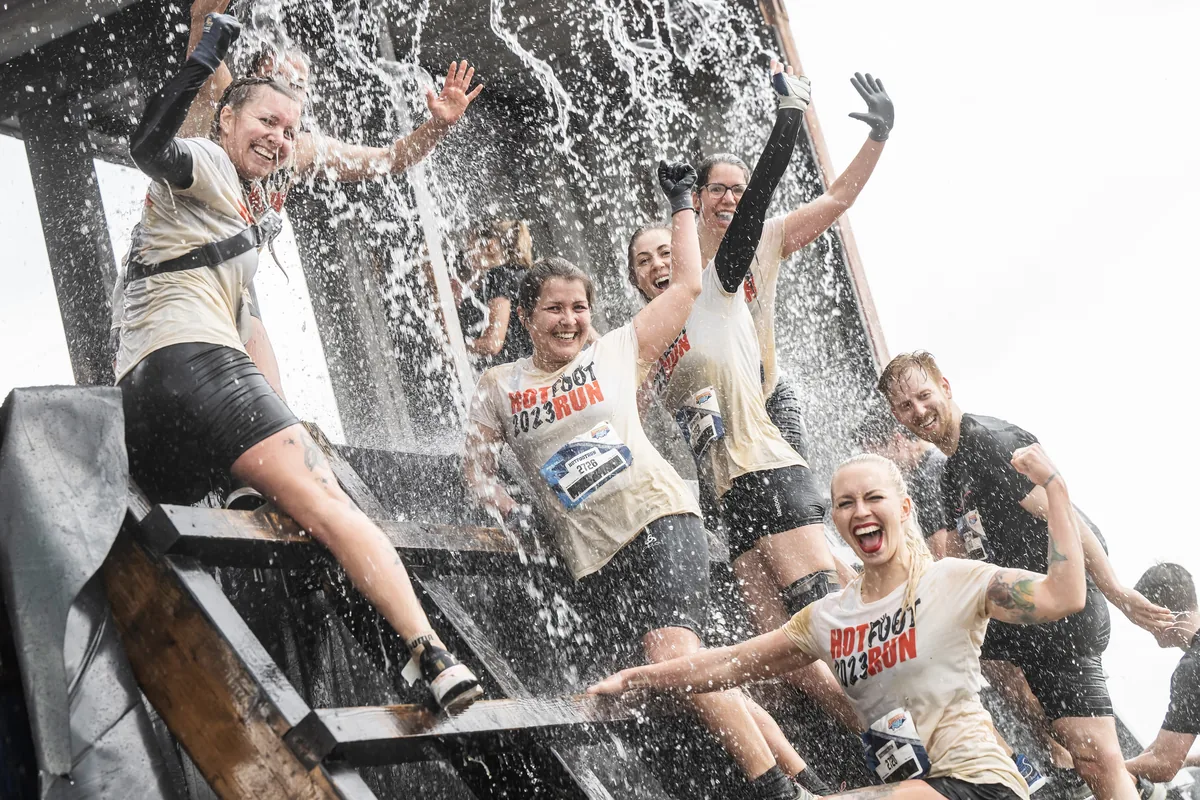 Läuferinnen und Läufer lachen unter einem Wasserhindernis beim Hotfoot Run